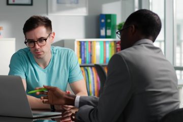 man pointing to a laptop while another man is looking on in an office setting