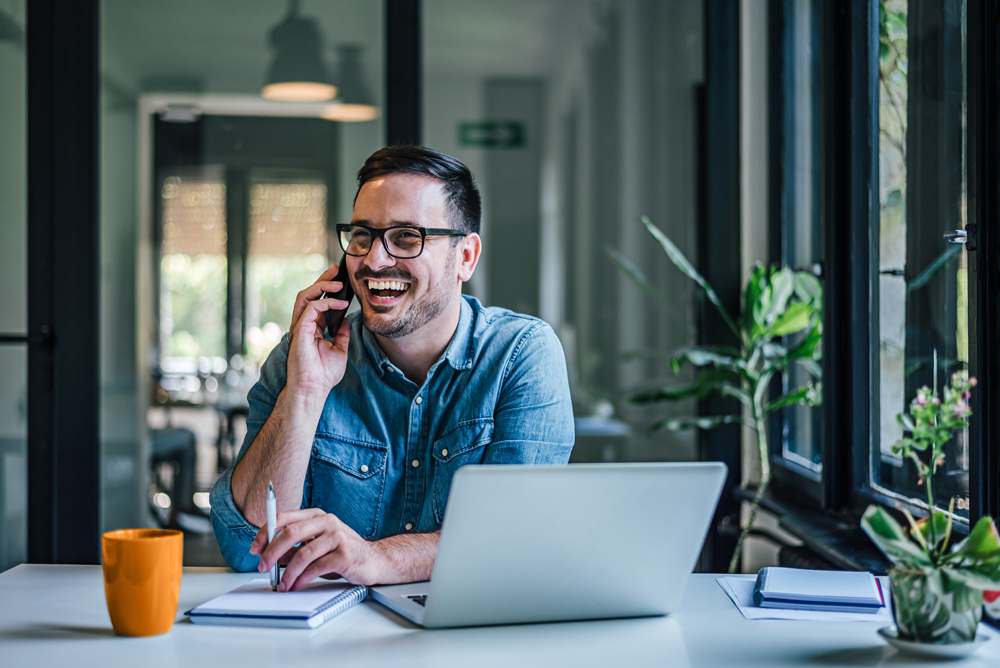 Man talking and smiling on a cell phone in front of a laptop on his desk in office setting