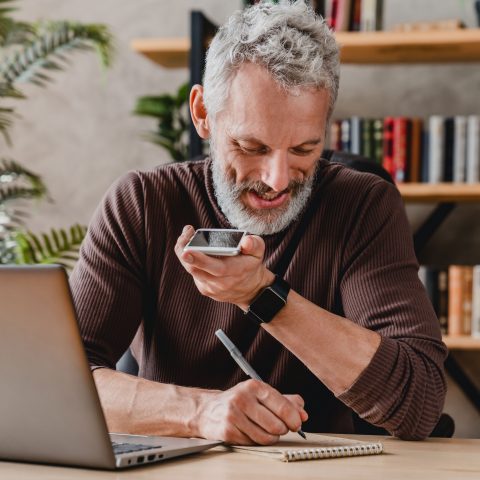 Older man with grey hair talking into a cell phone holding it near his mouth and writing notes on a pad of paper.